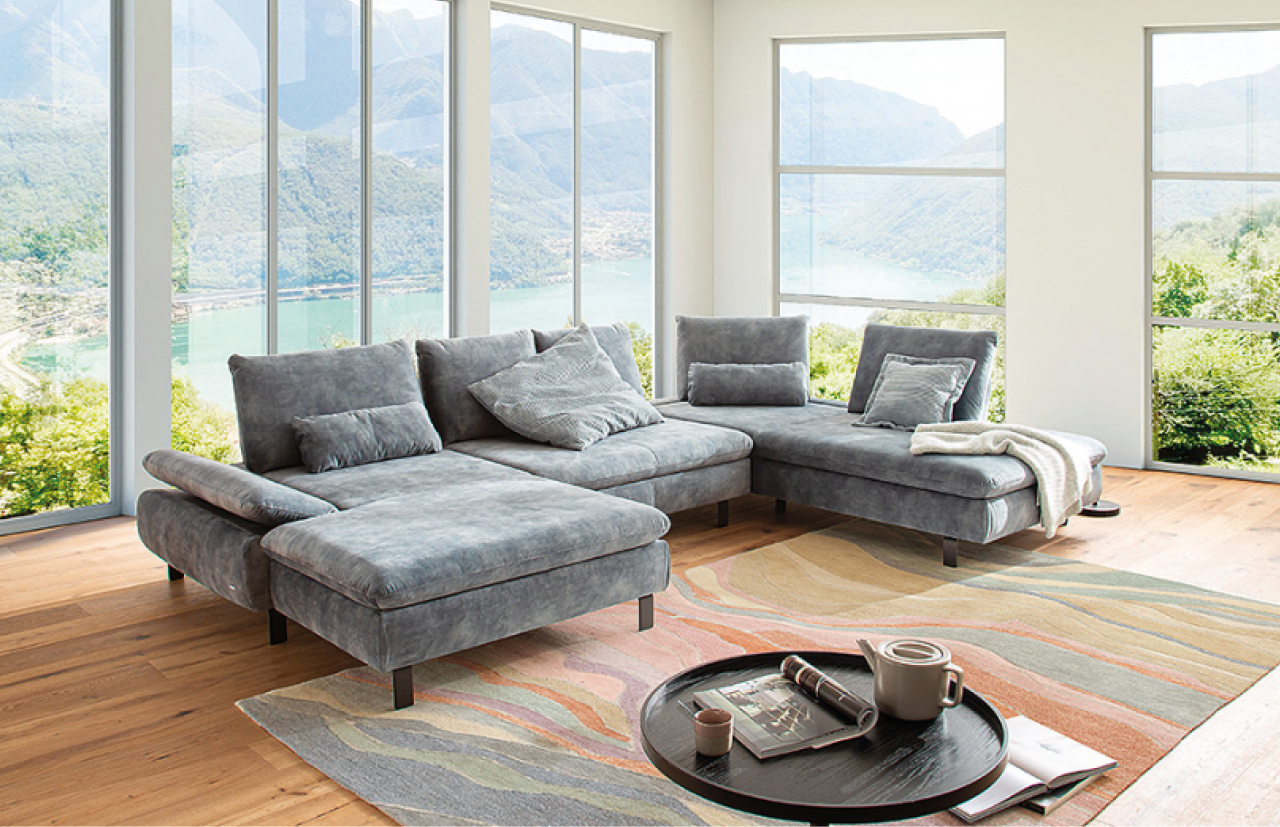 Graues Ecksofa mit passenden Kissen vor grosser Fensterfront in Wohnzimmer.