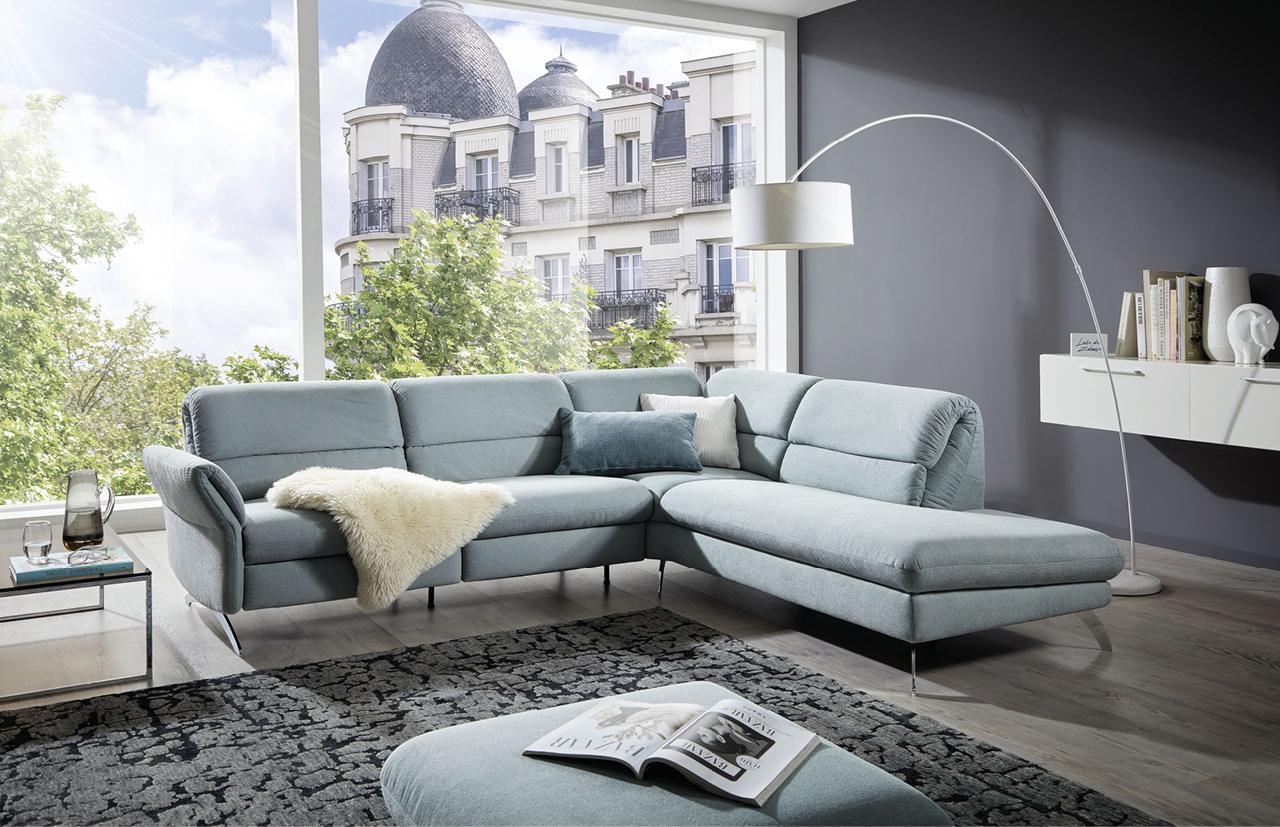 Großes Ecksofa in hellgrauem Stoff mit kuscheligem Fell, Zierkissen und passendem Hocker in elegantem Wohnzimmer mit Panoramafenster, Designer-Bogenlampe und modernem Teppich.