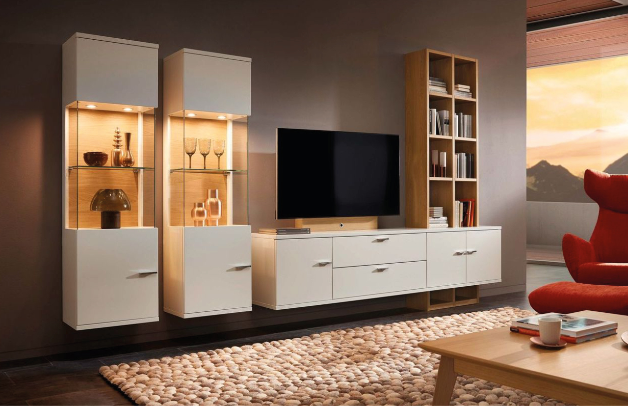 Stilvolle Wohnwand mit beleuchteten Vitrinen, TV-Board und Bücherregal in Weiß mit Holzelementen, dekoriert mit Gläsern und Vasen, ergänzt durch roten Sessel und hellen Teppich im Wohnzimmer.