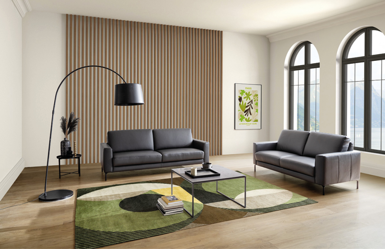 Helles Wohnzimmer mit zwei schwarzen Ledersofas, modernem grün-gemusterten Teppich, filigranem Couchtisch und schwarzer Bogenlampe vor großen Rundbogenfenstern und Holzpaneelwand.