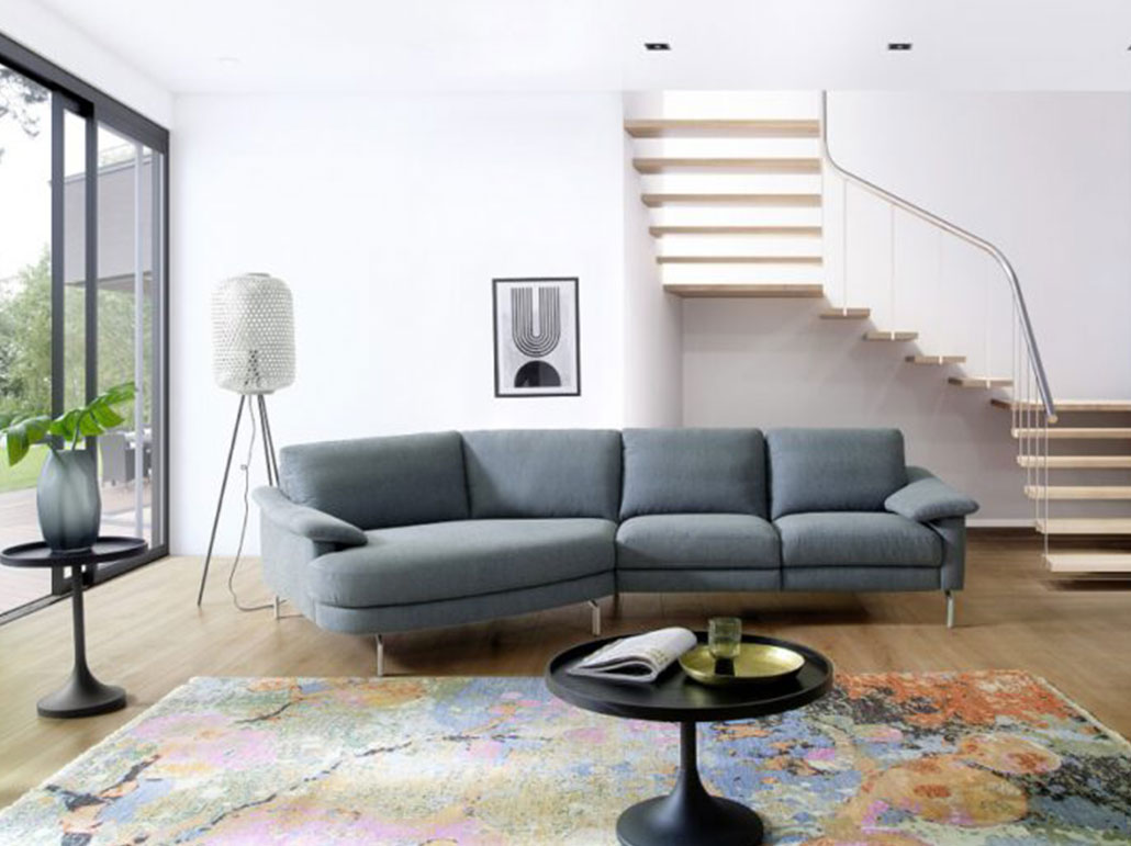 Modernes Ecksofa in Blau – stilvolle Wohnzimmergestaltung mit Designer-Flair Helles Wohnzimmer mit modernem blauen Ecksofa, bunter Designteppich, runder Couchtisch und offener Treppe – stilvolle Sofaecke, skandinavisches Interieur, großzügige Raumgestaltung mit Designmöbeln und klarer Linienführung.